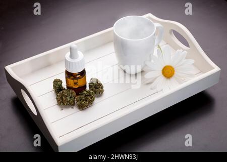 distributore di gocce d'olio di germogli marijuana e tazza bianca, fiore a margherita, su sfondo bianco di legno scuro vassoio Foto Stock