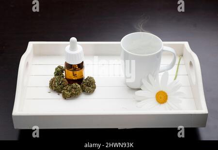 distributore di gocce d'olio di germogli marijuana e tazza bianca, fiore a margherita, su sfondo bianco di legno scuro vassoio Foto Stock