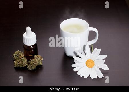boccioli di marijuana con flacone dosatore gocce, tazza bianca con ornamento di fiori a margherita per infusione di vapore, su tavola marrone, sfondo in legno Foto Stock