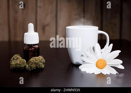 boccioli di marijuana con flacone dosatore gocce, tazza bianca con ornamento di fiori a margherita per infusione di vapore, su tavola marrone, sfondo in legno Foto Stock