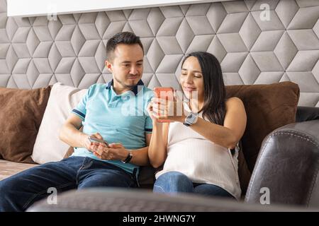 Coppia di uomo e donna incinta sorridendo usando il loro smartphone nel soggiorno della loro casa Foto Stock