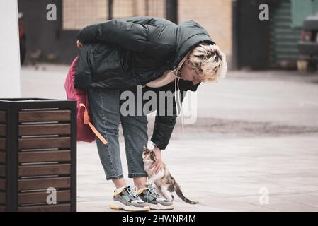 una ragazza in abiti casual si erge sul marciapiede e batte un gatto vagabo. Foto Stock