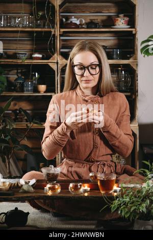 Donne che preparano la cerimonia del tè in stile boho ambiente con piante in studio. Tazzine trasparenti con bevanda calda in mano. Aroma di tradizionale Foto Stock