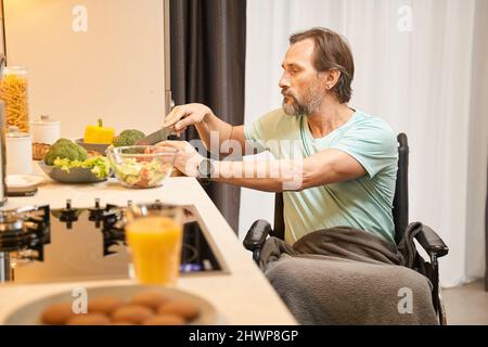 Uomo adulto in sedia a rotelle taglia le verdure in cucina Foto Stock