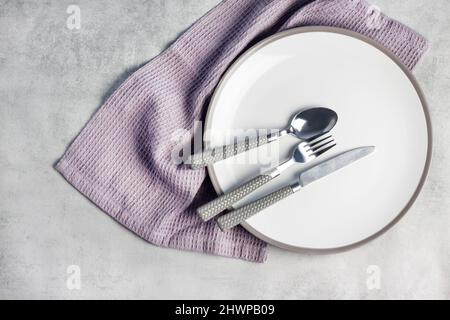 Impostazione del tavolo. Piatto bianco, posate e tovagliolo su sfondo grigio chiaro. Disposizione piatta, vista dall'alto. Foto Stock