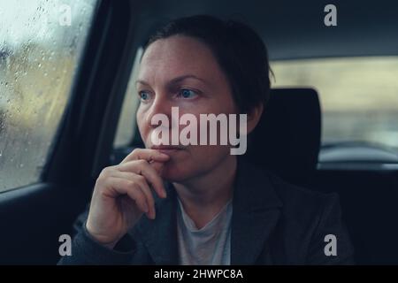 Donna d'affari delusa e preoccupata che guarda fuori la finestra del veicolo del taxi su freddo giorno piovoso sul suo modo di lavorare, fuoco selettivo Foto Stock