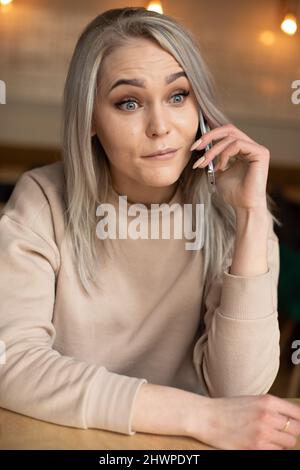 Foto verticale di sorpresa puzzled donna bionda pazza, fissando, comunicando il telefono cellulare in caffetteria, tavolo da seduta. Grandi cattive notizie Foto Stock