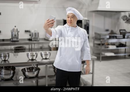 Bel cuoco asiatico cuoco in uniforme bianca scatta foto selfie al telefono o fa video blog per i social network mentre si lavora nella cucina del ristorante. Concetto di attività sociale sul posto di lavoro Foto Stock