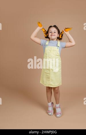 Ragazza allegra piccola che tocca trecce gialle kanekalon con le mani, saltando e giocando guardando la macchina fotografica con sorriso sul viso indossando tuta gialla e. Foto Stock