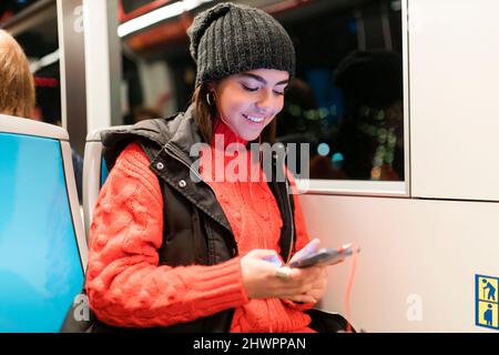 Sorridente giovane donna che usa lo smartphone e ascolta la musica con le cuffie intrauricolari in tram Foto Stock