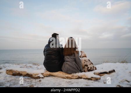 Giovane coppia seduta con il cane Pastore Australiano su tronchi coperti di neve in spiaggia Foto Stock