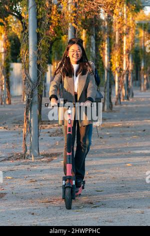 Donna d'affari sorridente che guida scooter elettrico push con uomo d'affari dietro di lei su strada Foto Stock