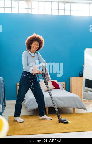 Sorridente Afro giovane donna in piedi con aspirapolvere su tappeto in camera da letto a casa Foto Stock