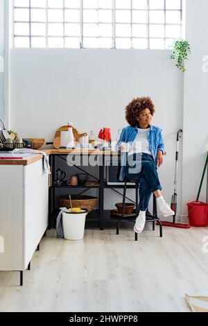 Giovane donna afro pensierosa seduta su sgabello appoggiata sul bancone della cucina rilassante a casa Foto Stock