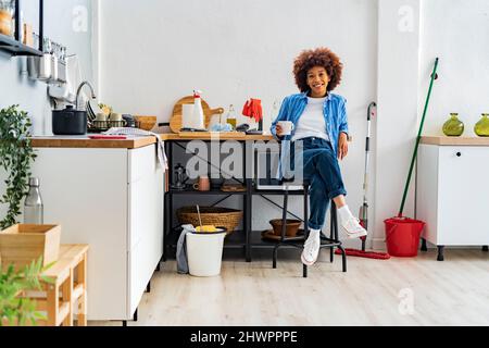 Sorridente giovane donna afroica seduta su sgabello appoggiata sul bancone della cucina rilassarsi a casa Foto Stock