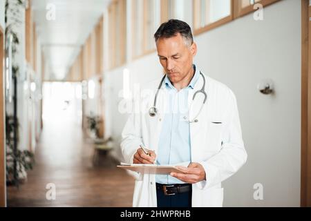 Medico maturo che scrive la cartella medica nel corridoio in ospedale Foto Stock