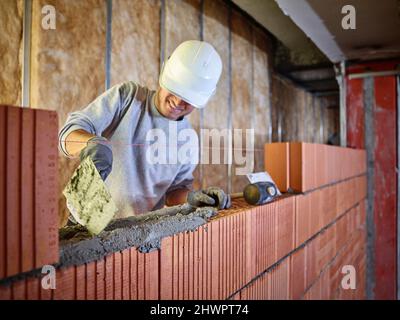 Muratore sorridente che applica cemento su mattoni che lavorano in cantiere Foto Stock