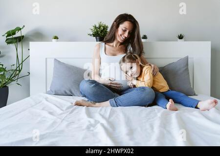Donna incinta felice seduta con la figlia a letto a casa Foto Stock