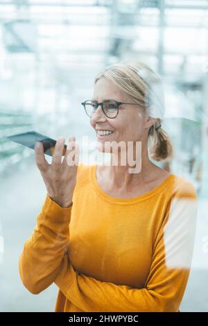 Donna d'affari che invia la posta vocale sul telefono cellulare visto attraverso il vetro dell'ufficio Foto Stock