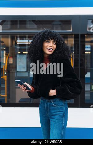 Giovane donna sorridente con smartphone di fronte al tram Foto Stock