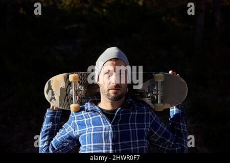 Uomo che indossa un cappello a maglia che porta lo skateboard a spalla Foto Stock