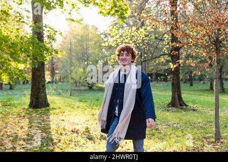Uomo sorridente con capelli ricci rossi e sciarpa in piedi nel parco autunnale Foto Stock