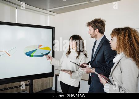 Giovane donna d'affari sorridente che spiega il grafico a torta ai colleghi in ufficio Foto Stock