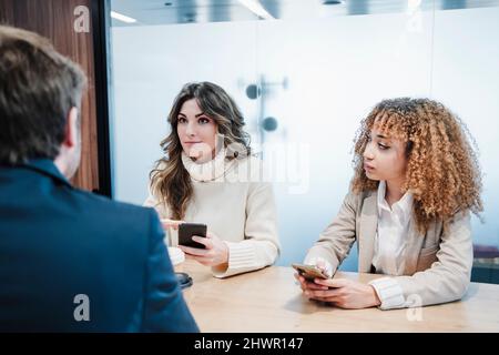 Donne d'affari con smartphone che parlano con uomo d'affari seduto alla scrivania Foto Stock