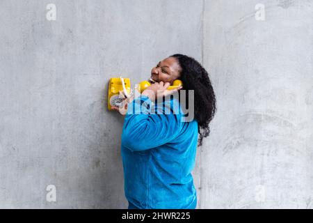 Donna allegra che parla attraverso il ricevitore telefonico vecchio stile di fronte al muro grigio Foto Stock