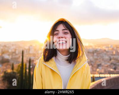 Giovane donna sorridente in impermeabile giallo con la città sullo sfondo al tramonto Foto Stock