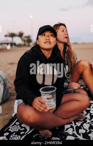 Donna premurosa con una tazza di birra seduta da un amico in spiaggia Foto Stock