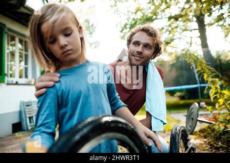 Uomo sorridente che guarda il figlio che pulisce la bicicletta nel cortile Foto Stock