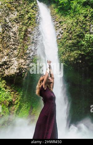 Donna in piedi con le braccia sollevate di fronte alla cascata la Fortuna, Costa Rica Foto Stock