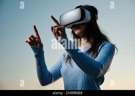 Giovane donna che utilizza simulatore di realtà virtuale gesturando di fronte al cielo del tramonto Foto Stock