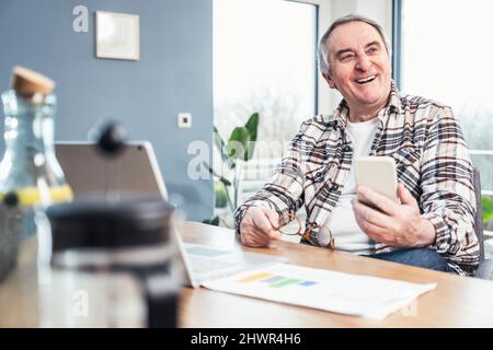 Uomo anziano allegro che tiene il telefono intelligente e gli occhiali seduti al tavolo Foto Stock