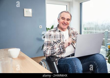Uomo felice con un computer portatile seduto su sedia a rotelle a casa Foto Stock