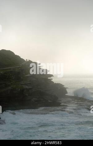 Una persona cammina lungo un sentiero in cima alla scogliera accanto alle grandi onde dell'oceano che si infrangono sulla costa rocciosa in una mattinata di nebbia a Bronte Beach, Sydney, Australia Foto Stock