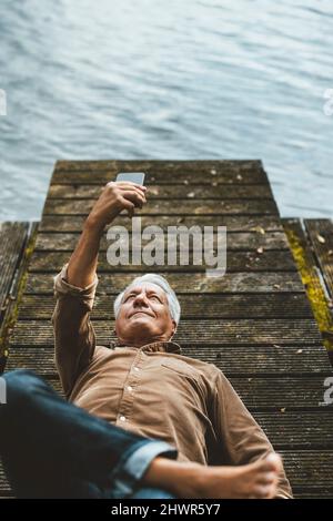Uomo anziano che prende selfie attraverso lo smart phone che giace sul jetty vicino al lago Foto Stock