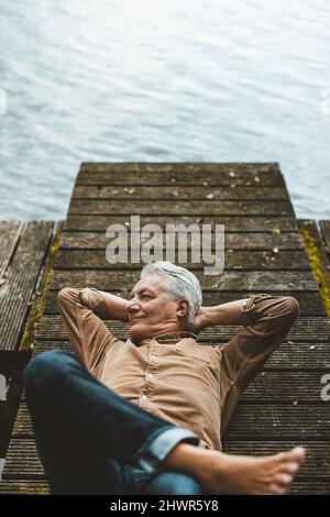 Uomo anziano sorridente con le mani dietro la testa adagiato sul molo vicino al lago Foto Stock