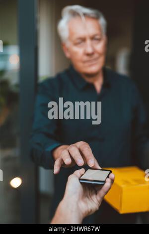 Uomo anziano con la firma del pacchetto sullo smartphone tenuto dal responsabile della consegna Foto Stock