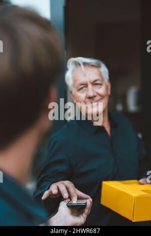 Uomo anziano sorridente con firma pacchetto giallo sul telefono cellulare tenuto dalla persona di consegna Foto Stock