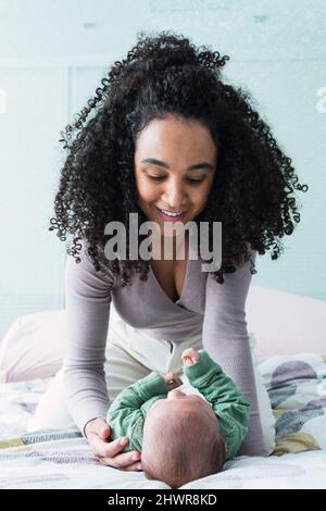 Madre sorridente che guarda il bambino che si trova sul letto di casa Foto Stock