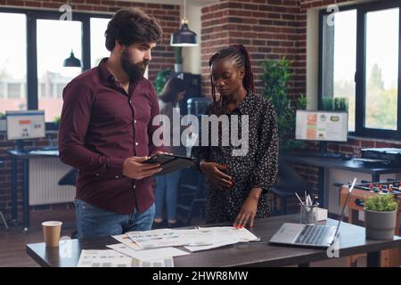 Collaboratore sul luogo di lavoro che sottolinea sul tablet che la contabilità ha dichiarato entrate aziendali errate. Consulente finanziario incinta afroamericano che discute con il collega circa la rappresentazione errata del grafico. Foto Stock