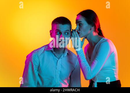 Ritratto di due giovani, donna che sussurra sull'orecchio dell'uomo, notizia parlante isolata su sfondo arancione in luci al neon Foto Stock