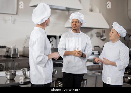 Tre cuochi chef multirazziale parlano mentre si levano in piedi insieme durante una pausa nella cucina del ristorante. Chef asiatici, latini ed europei che indossano una divisa bianca al lavoro Foto Stock