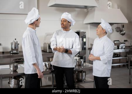 Tre cuochi chef multirazziale parlano mentre si levano in piedi insieme durante una pausa nella cucina del ristorante. Chef asiatici, latini ed europei che indossano una divisa bianca al lavoro Foto Stock