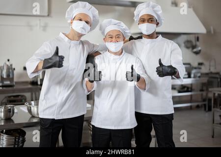 Ritratto di squadra multirazziale di tre chef in piedi insieme e mostrando OK segno. Chef ben vestiti che indossano maschere per il viso e guanti protettivi per prevenire la diffusione di infezioni durante la pandemia Foto Stock