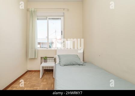 Letto singolo con copriletto blu con testiera bianca, tavolo laterale abbinato e finestra con tende bianche in una camera da letto con pavimento in parquet Foto Stock