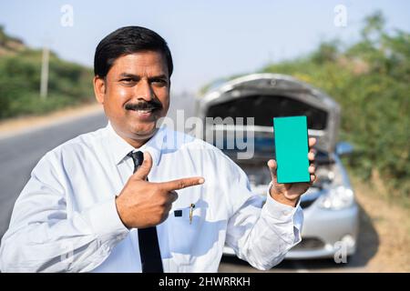 Felice uomo d'affari che mostra il telefono cellulare con schermo verde di fronte alla macchina rotta guardando la macchina fotografica - concetto di prenotazione online di servizio di riparazione auto Foto Stock