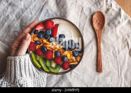 Colazione sana in ciotola di cocco sul tavolo con tovaglia di lino. Yogurt con fiocchi di mais e mirtillo, fragola, kiwi e lampone. Mano donne Foto Stock